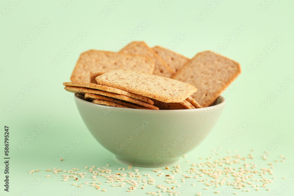 Bowl with tasty crackers and scattered sesame seeds on green background