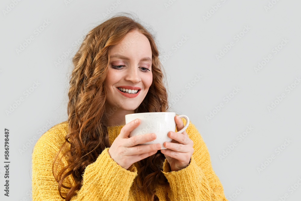 Beautiful redhead woman with cup of tea on grey background