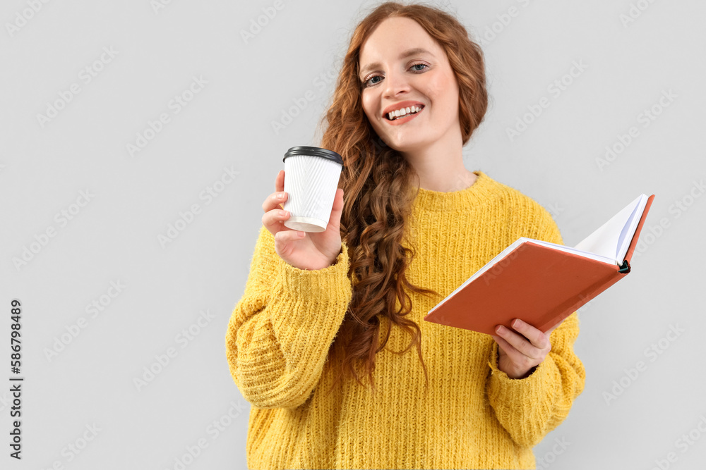 Beautiful redhead woman with cup of coffee and book on grey background