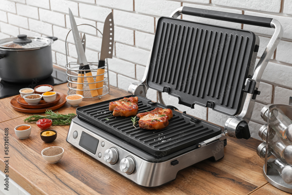 Modern electric grill with tasty steaks and spices on table near white brick wall