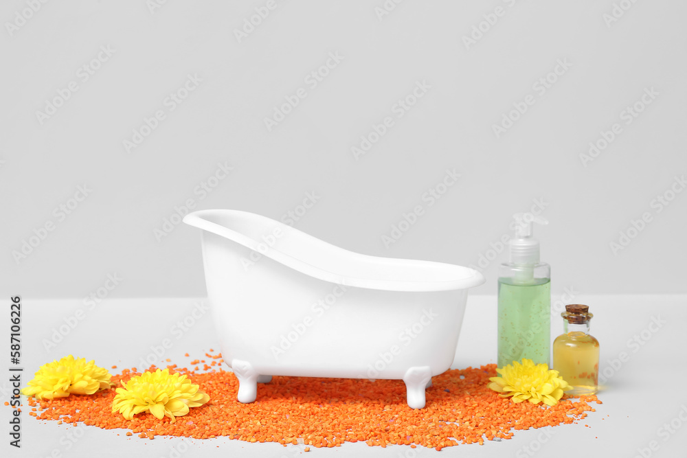 Small bathtub, bottles of cosmetic products, flowers and sea salt on light background