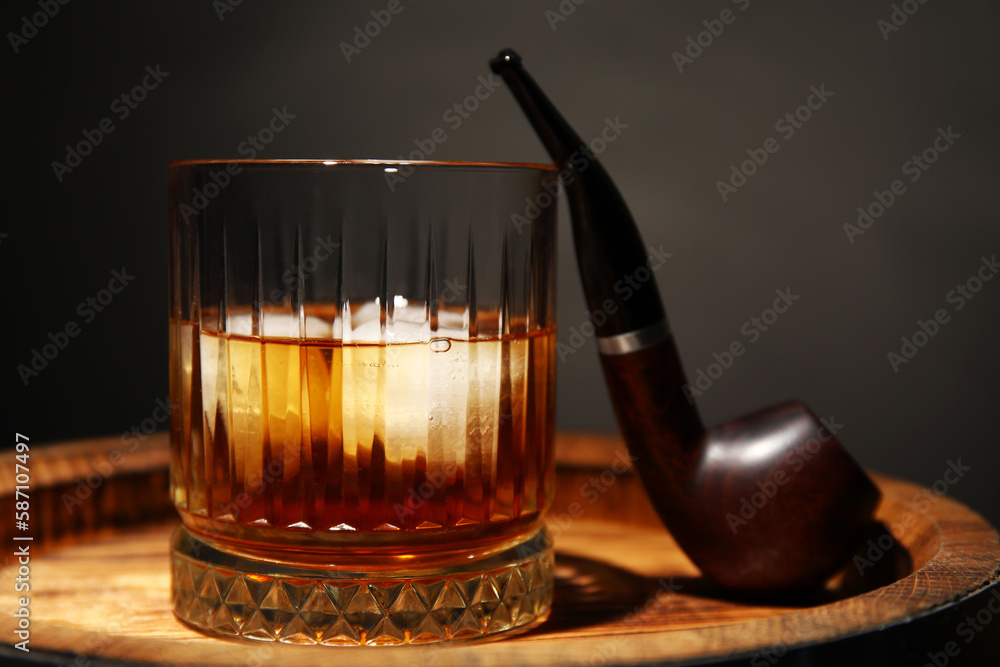 Wooden barrel with glass of cold rum and smoking pipe on dark background