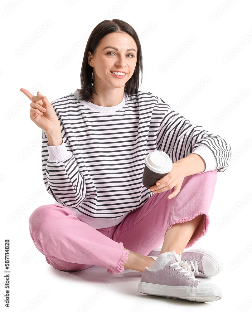 Pretty young woman with cup of hot coffee pointing at something against white background