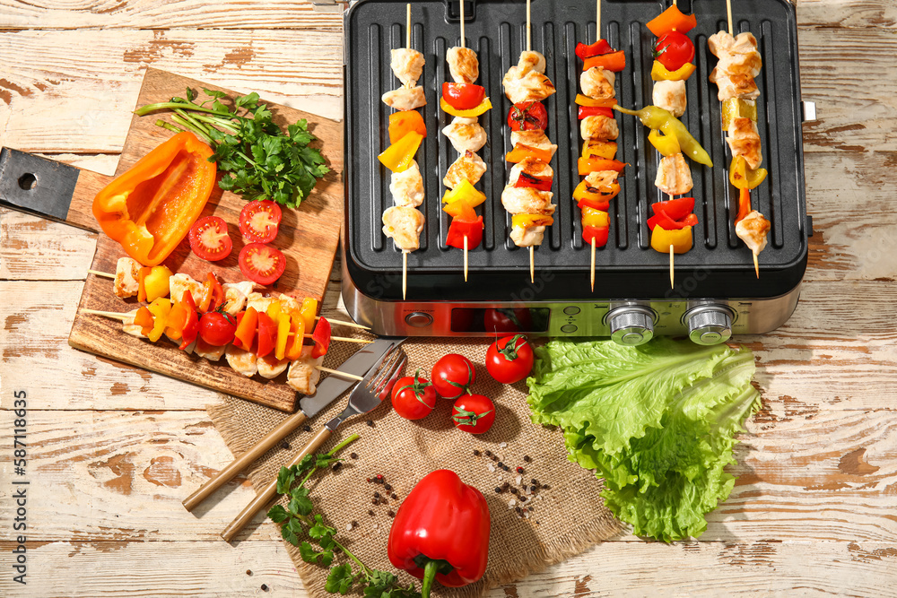 Modern electric grill with tasty chicken skewers and vegetables on light wooden background