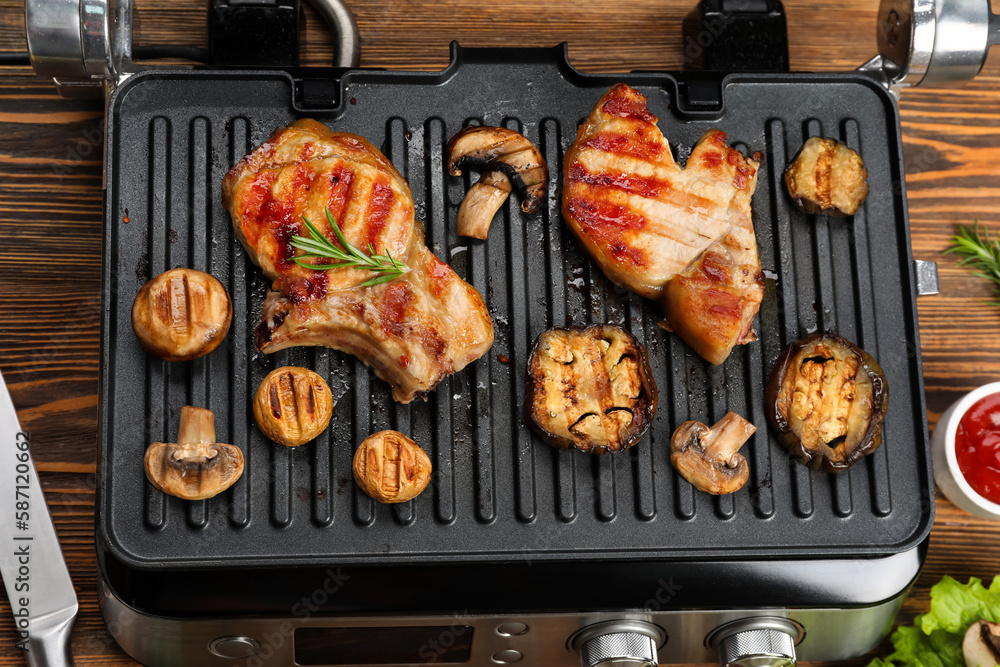 Modern electric grill with tasty steaks and vegetables on wooden background