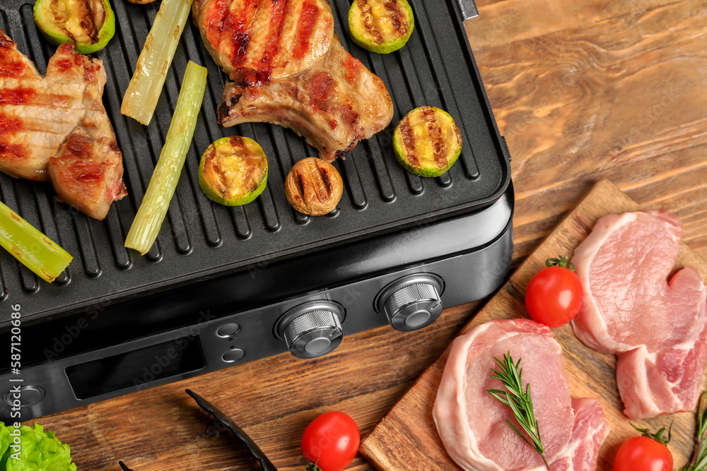 Modern electric grill with tasty steaks and vegetables on wooden background, closeup