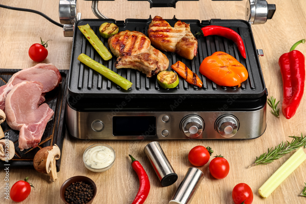 Modern electric grill with tasty steaks and vegetables on wooden background