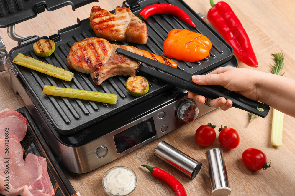 Woman cooking tasty steaks and vegetables on modern electric grill at table