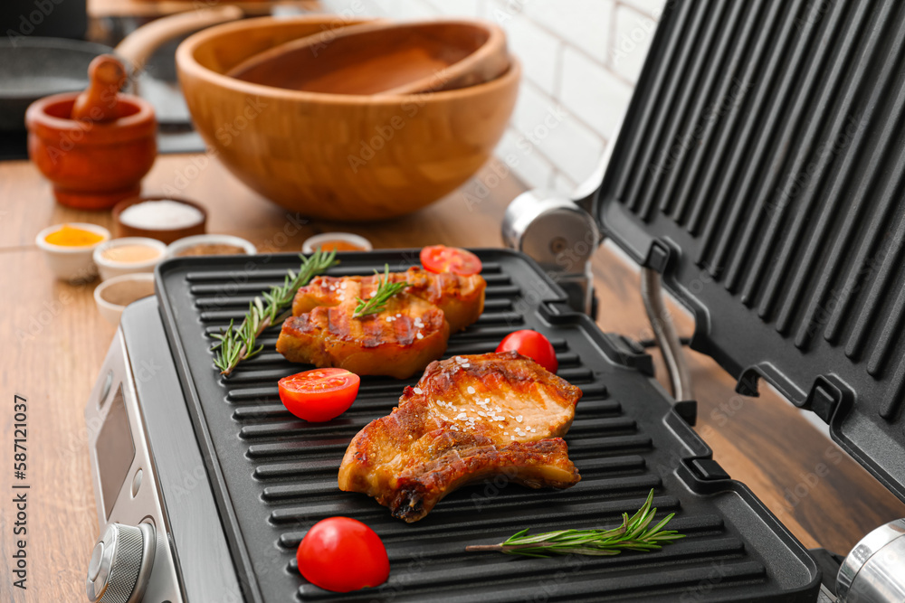 Modern electric grill with tasty steaks and tomatoes on table near white brick wall