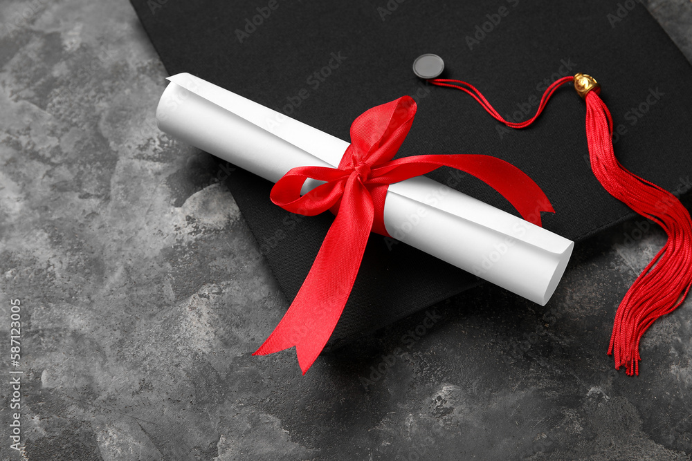 Diploma with red ribbon and graduation hat on dark grey table