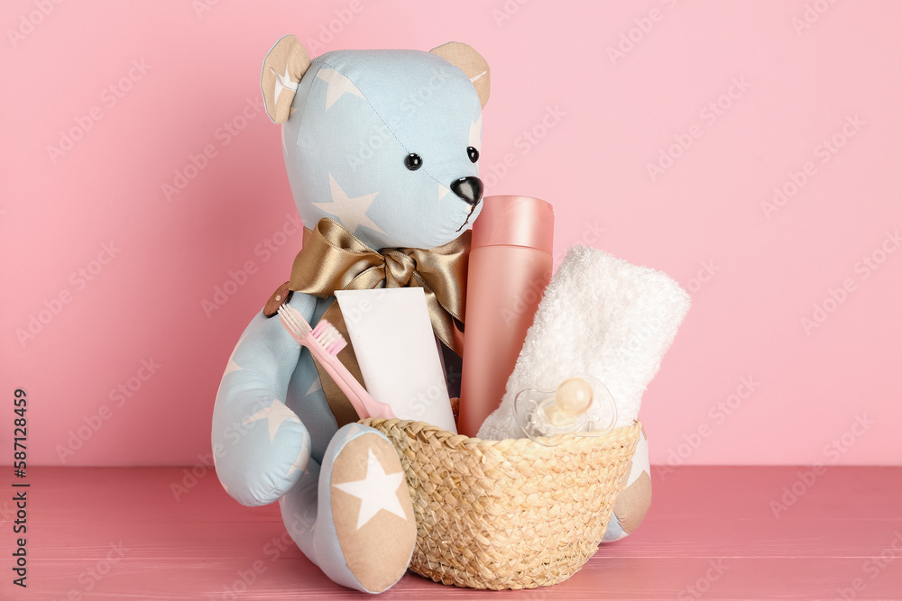 Toy bear and basket with baby accessories on table near pink wall