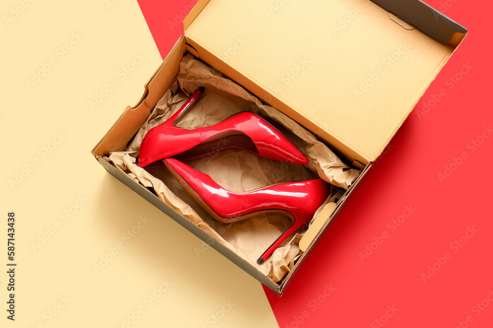 Cardboard box with high heeled shoes on color background