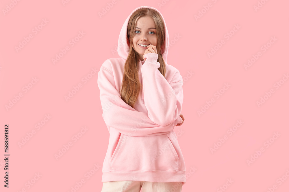 Young woman in hoodie biting nails on pink background