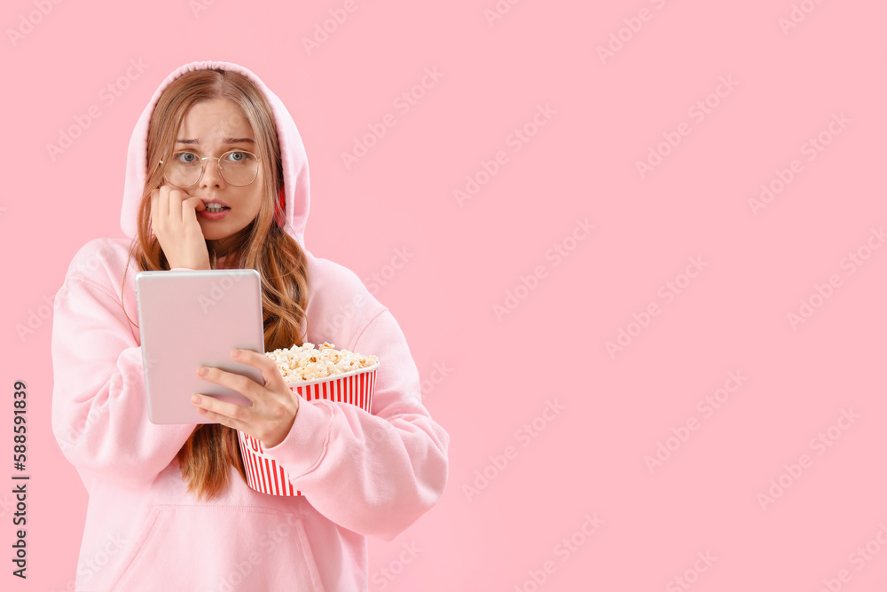 Young woman with tablet computer and popcorn biting nails on pink background