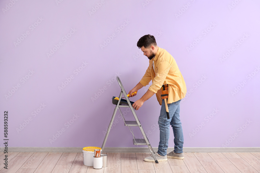 Young man with paint roller and ladder near lilac wall