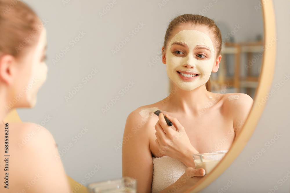 Young woman applying turmeric mask with brush near mirror at home