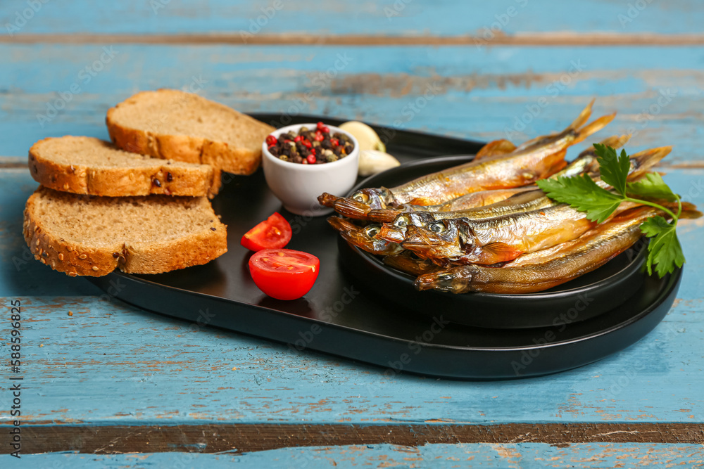 Board with tasty smoked capelin and bread pieces on blue wooden background