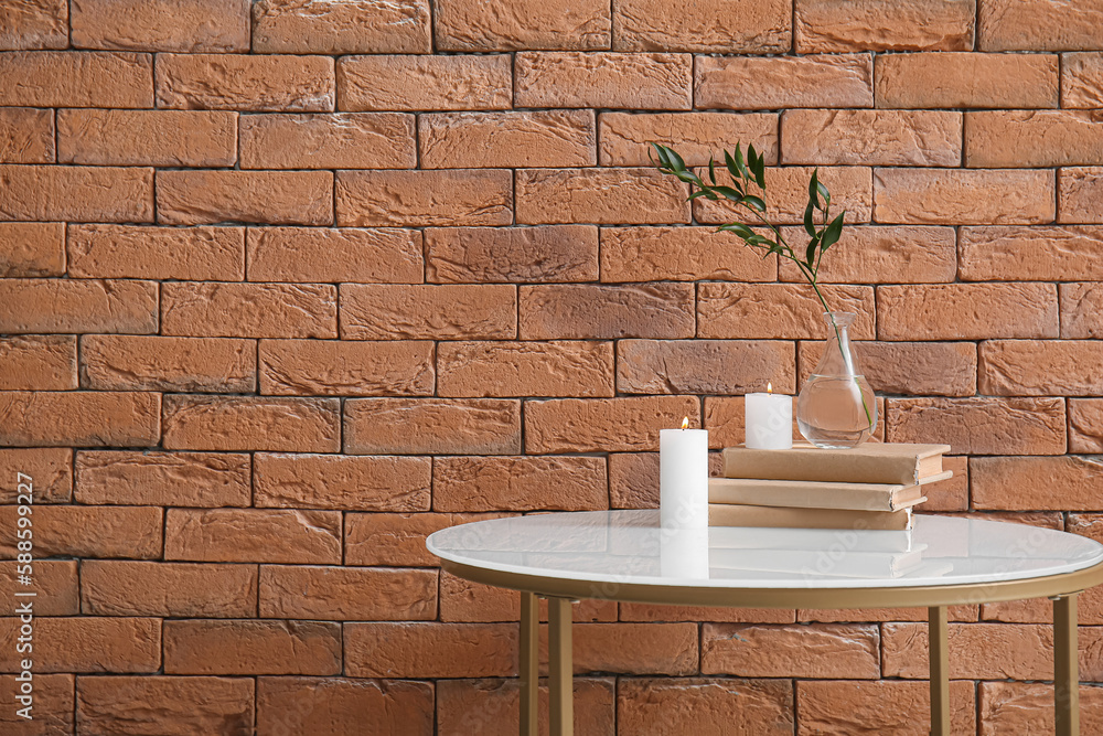 Burning candles, books and vase with ruscus branch on table near brown brick wall