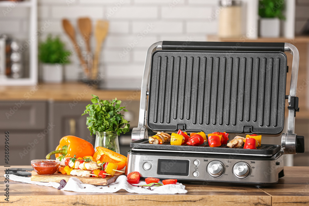 Modern electric grill with tasty chicken skewers and vegetables on table in kitchen