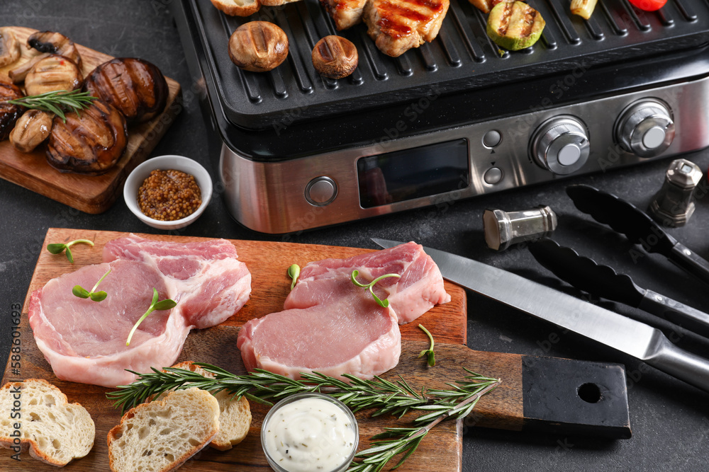 Wooden board with raw steaks and modern electric grill on dark background