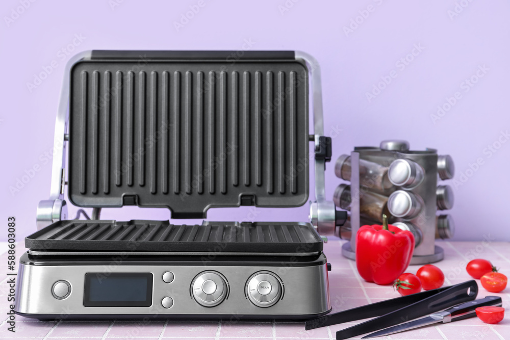 Modern electric grill with open lid, tongs and vegetables on pink tile table against lilac backgroun