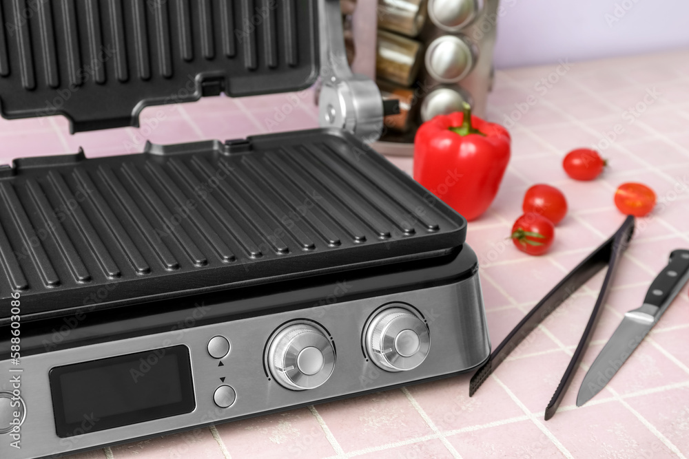 Modern electric grill with open lid, tongs and vegetables on pink tile table