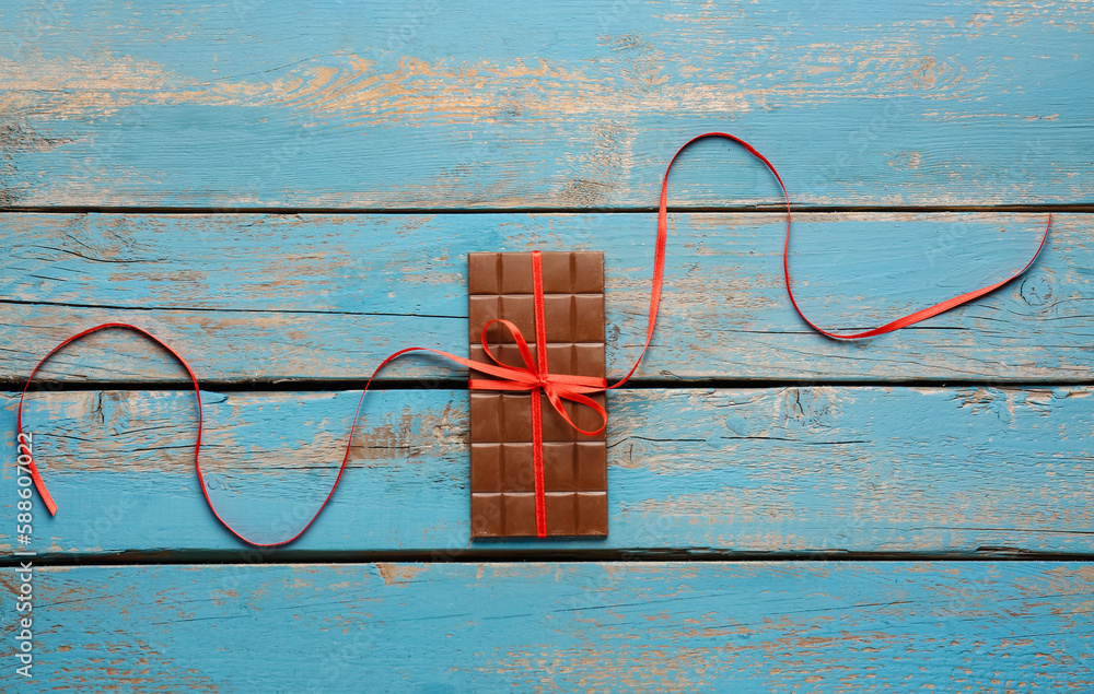 Tasty chocolate bar tied with red ribbon on color wooden background