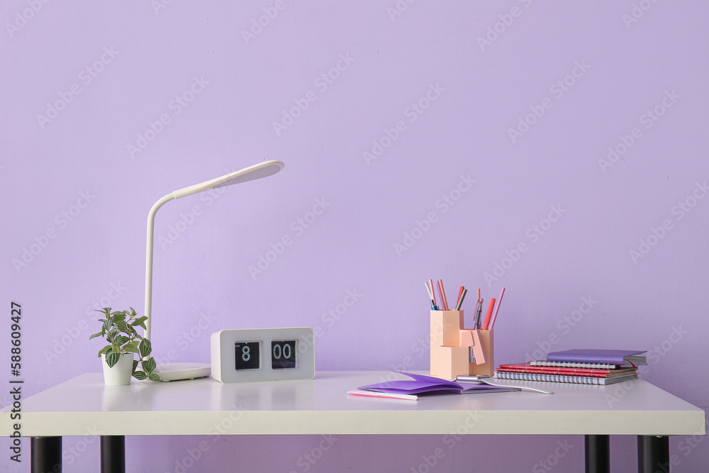 Workplace with stationery, clock and lamp near lilac wall