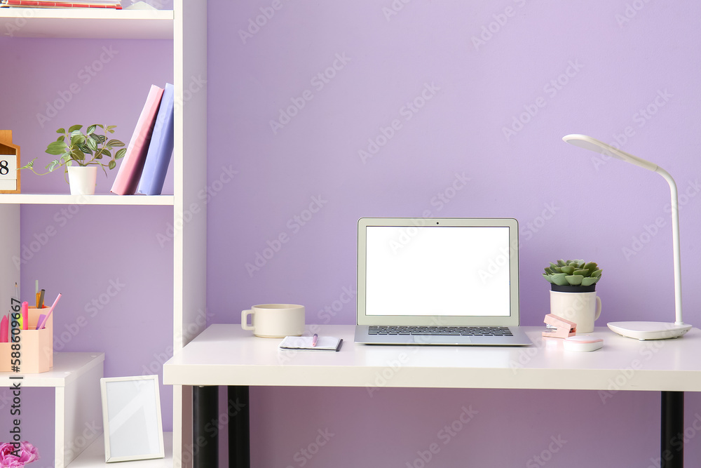 Workplace with laptop cup and stationery near lilac wall in office