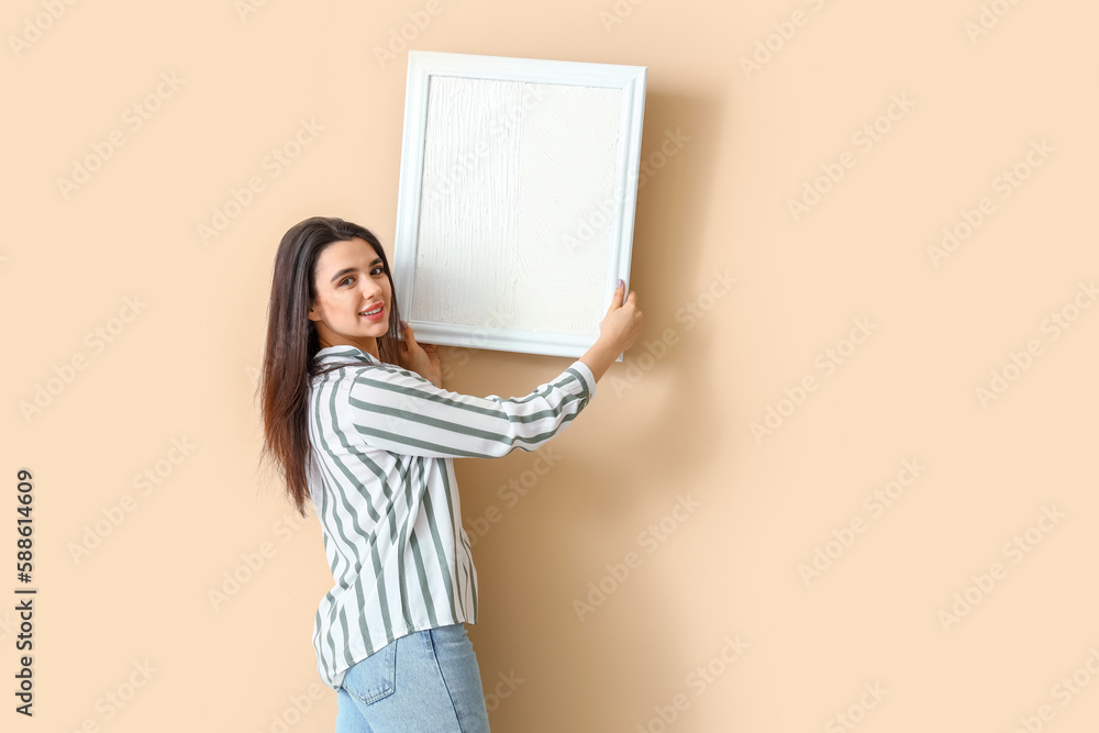 Young woman hanging blank picture on beige wall