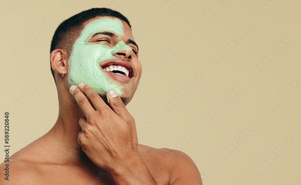 Male skincare routine: Happy young man smiling while pampering his skin with a face mask