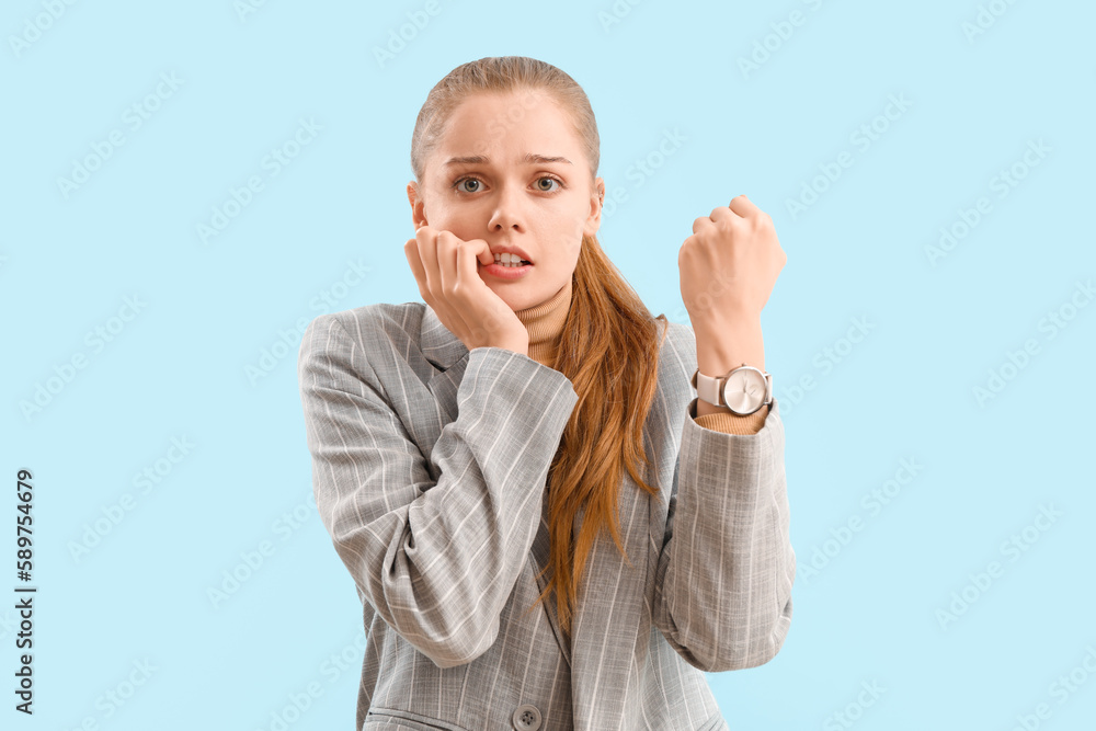 Young businesswoman with wristwatch biting nails on blue background