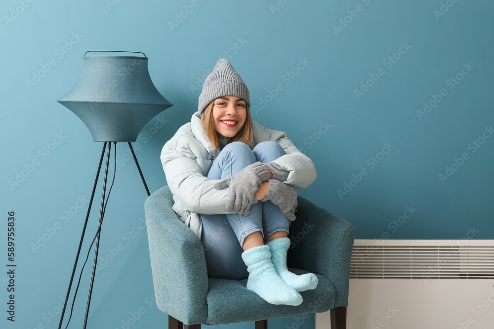 Frozen young woman in winter clothes heating near radiator at home