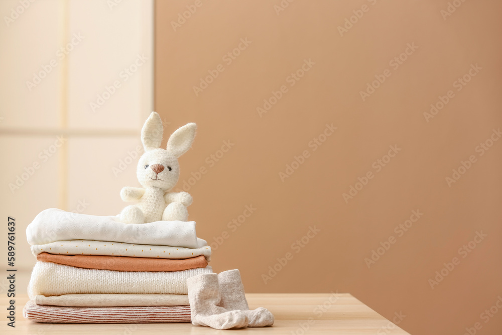 Stack of baby clothes, socks and toy bunny on table against color wall