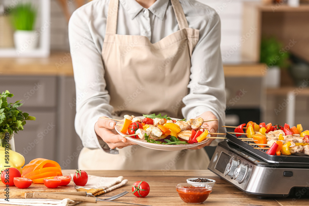 Woman holding plate with grilled chicken skewers and vegetables in kitchen, closeup