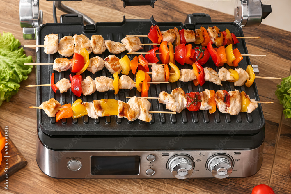 Modern electric grill with tasty chicken skewers and vegetables on table