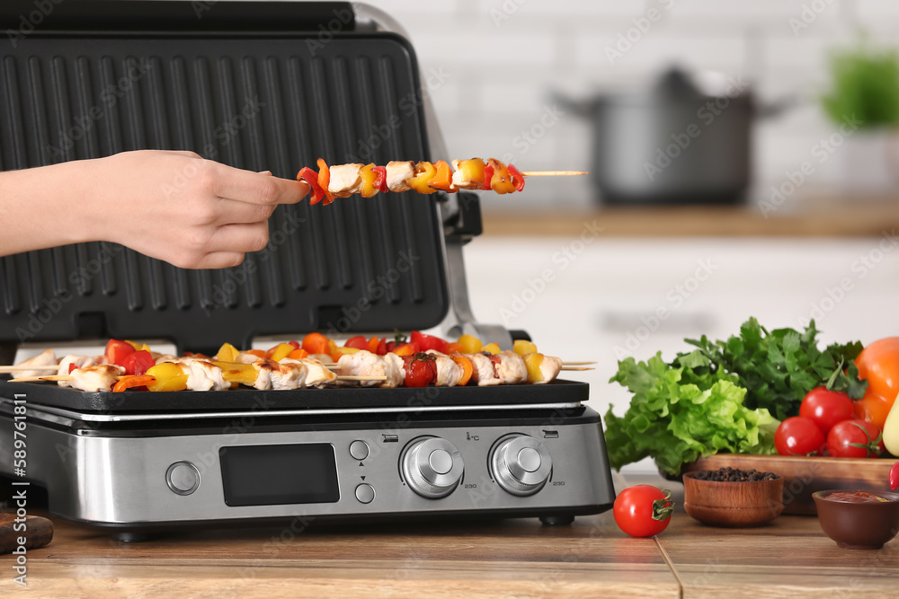 Woman cooking tasty chicken skewers and vegetables on modern electric grill at table in kitchen