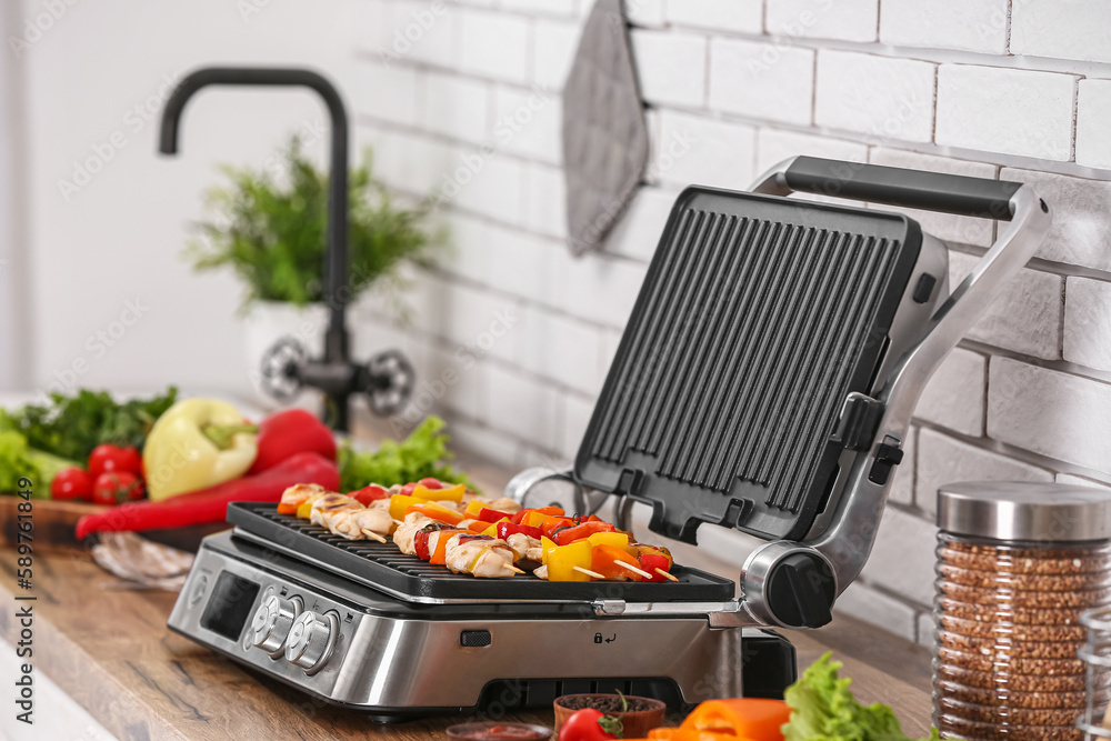 Modern electric grill with tasty chicken skewers and vegetables on table near light brick wall