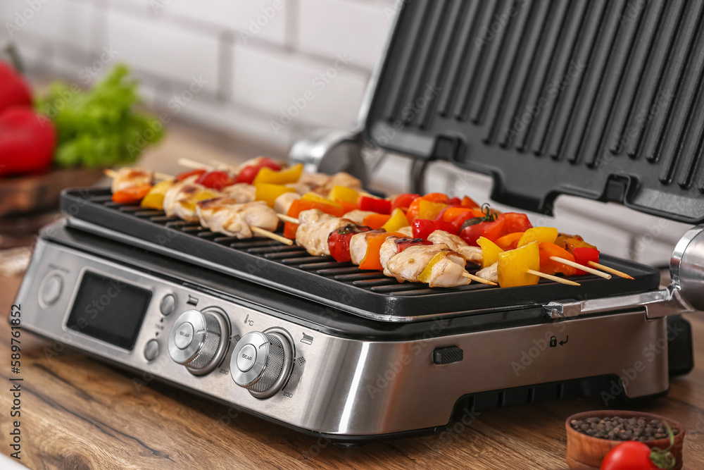 Modern electric grill with tasty chicken skewers and vegetables on table near light brick wall