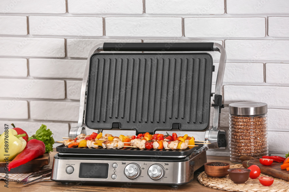 Modern electric grill with tasty chicken skewers and vegetables on table near light brick wall