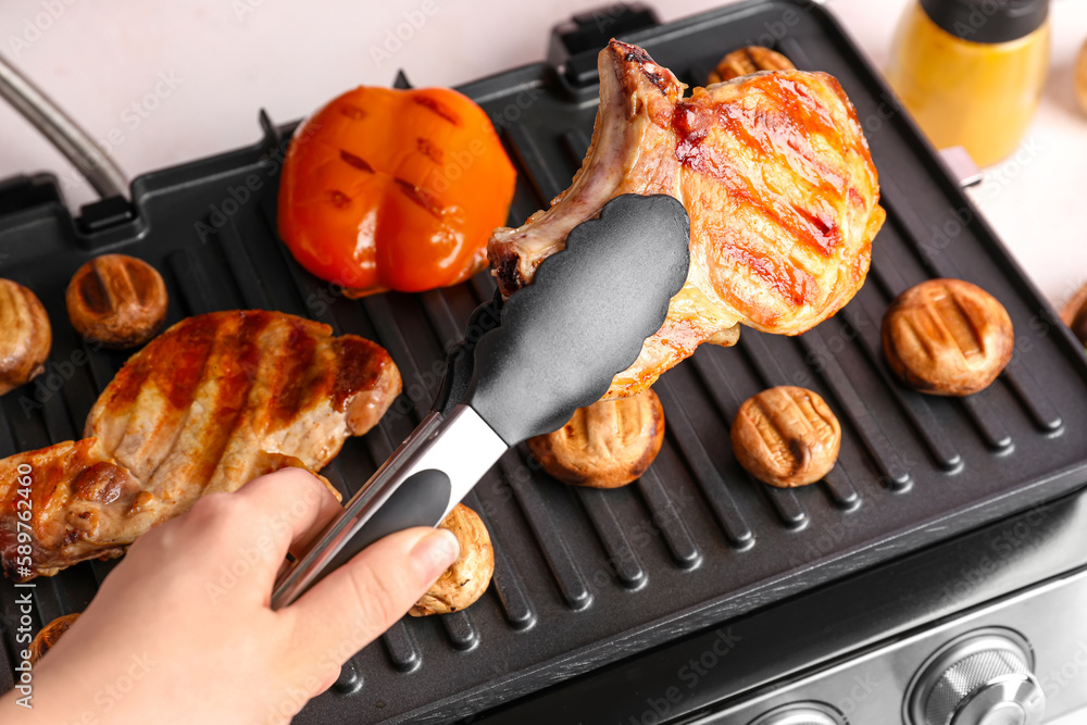 Woman cooking tasty steaks and vegetables on modern electric grill at table, closeup