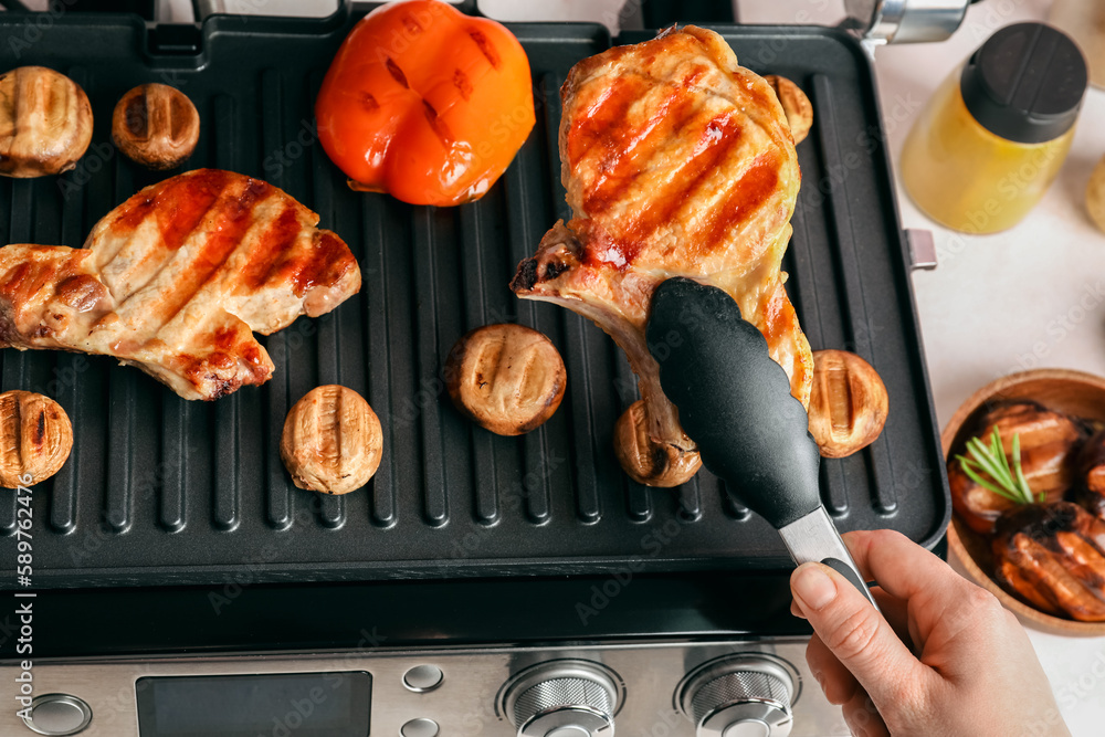 Woman cooking tasty steaks and vegetables on modern electric grill at table, closeup