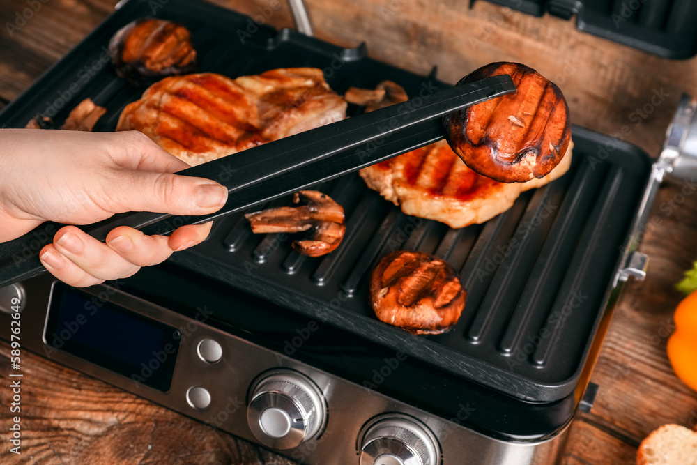 Woman cooking tasty steaks and vegetables on modern electric grill at table, closeup