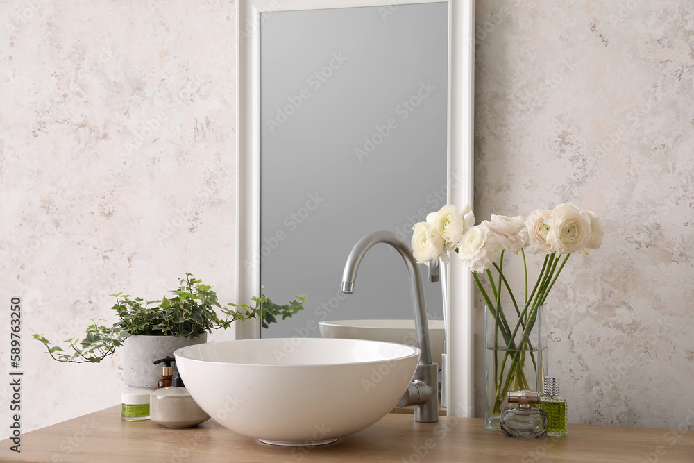 Vase with flowers, sink, mirror and bath accessories on counter in bathroom