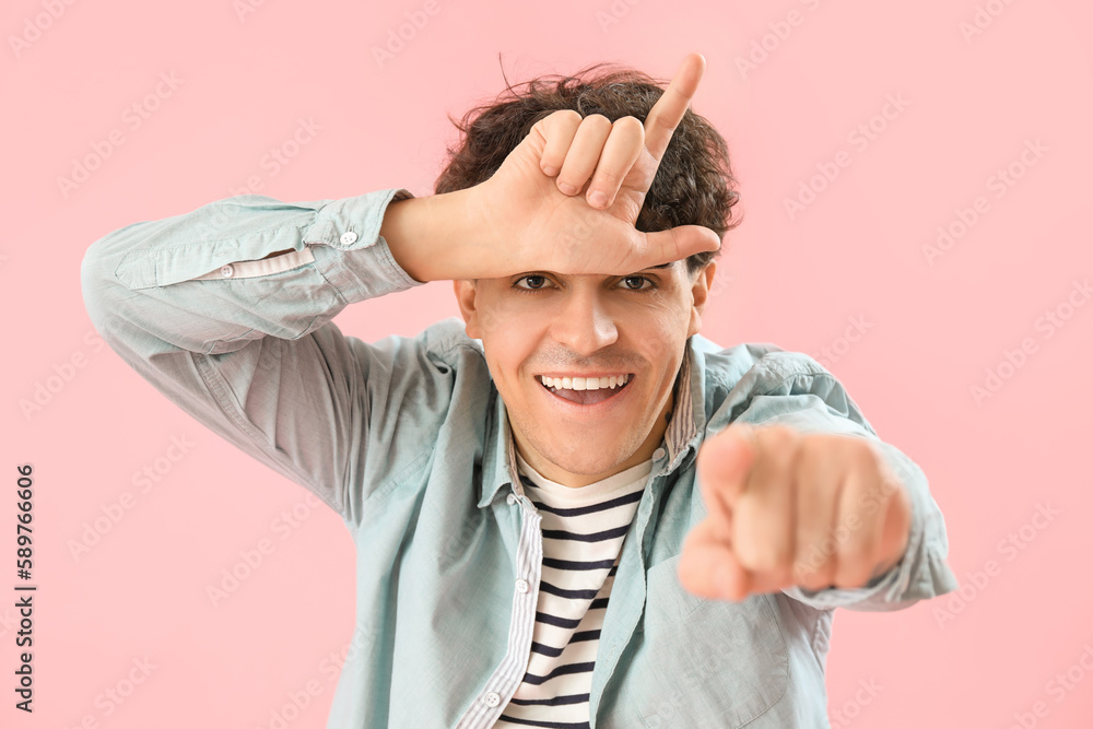 Laughing young man showing loser gesture and pointing at viewer on pink background