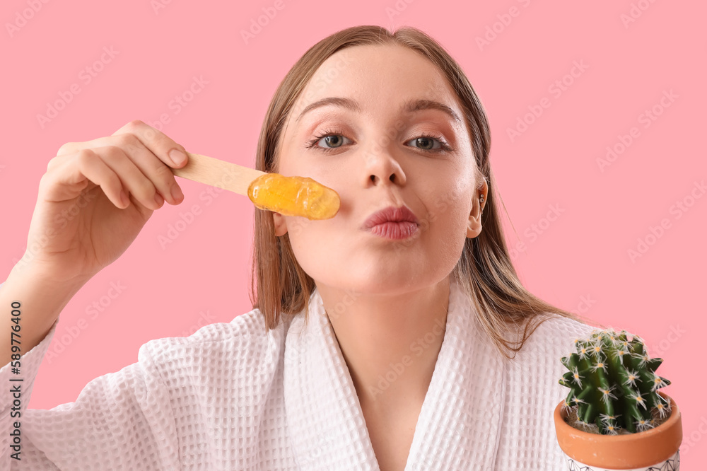 Young woman holding spatula with sugaring paste and cactus on pink background