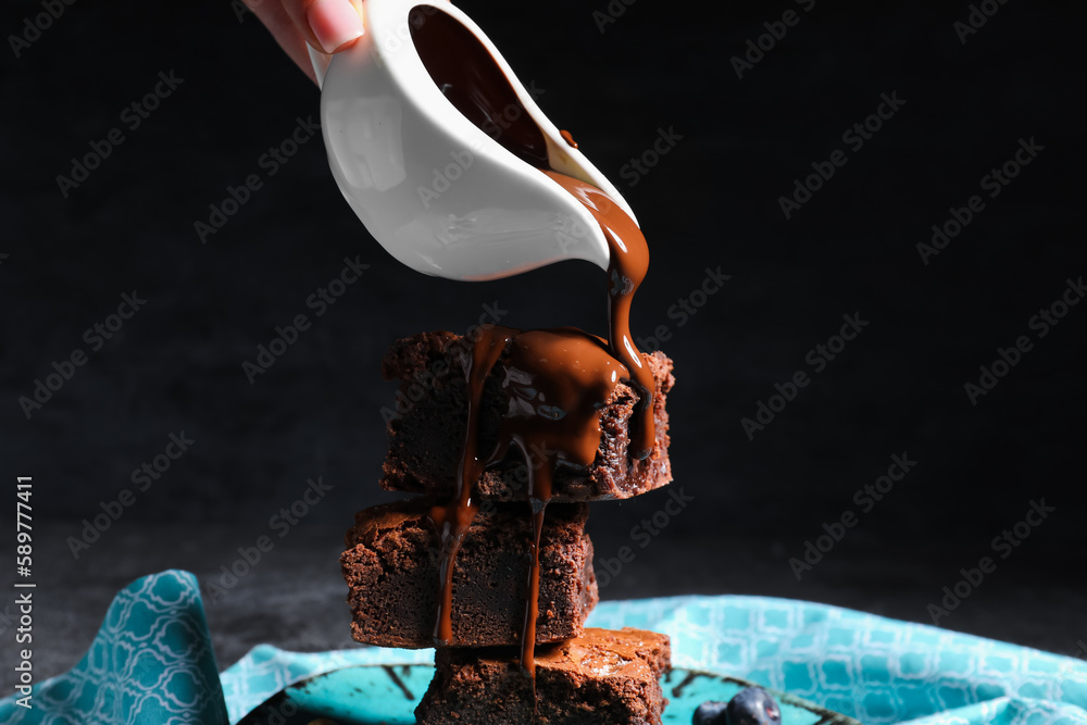 Woman pouring topping onto pieces of tasty chocolate brownie on black background