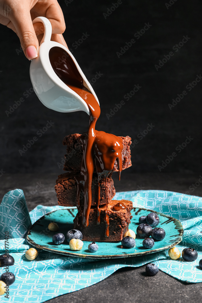 Woman pouring topping onto plate with pieces of tasty chocolate brownie on black background