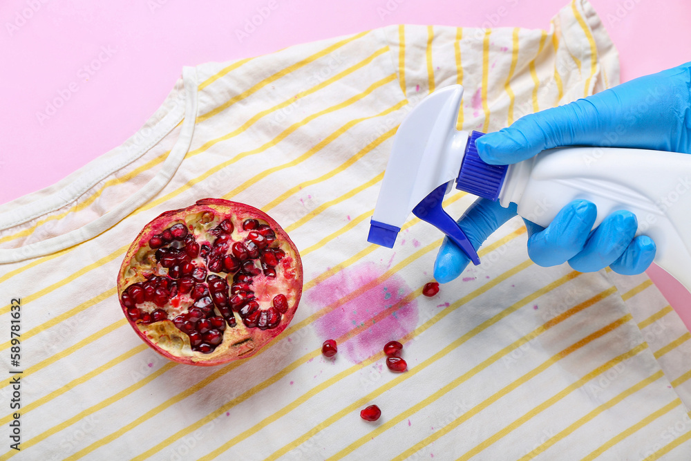 Woman spraying detergent onto stained t-shirt with pomegranate on pink background, closeup