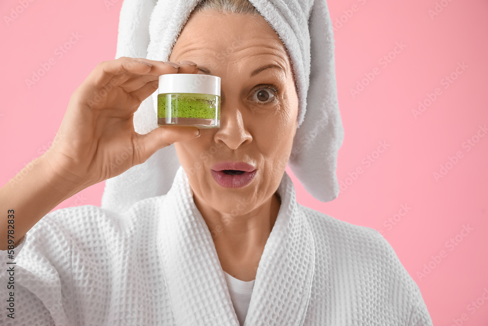 Surprised mature woman with cosmetic cream on pink background, closeup